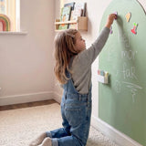 Meisje op knieën plaatst magneten op MagneetBoog+ in Noorderlicht (stofgroen) met krijttekst "hallo mama papa"